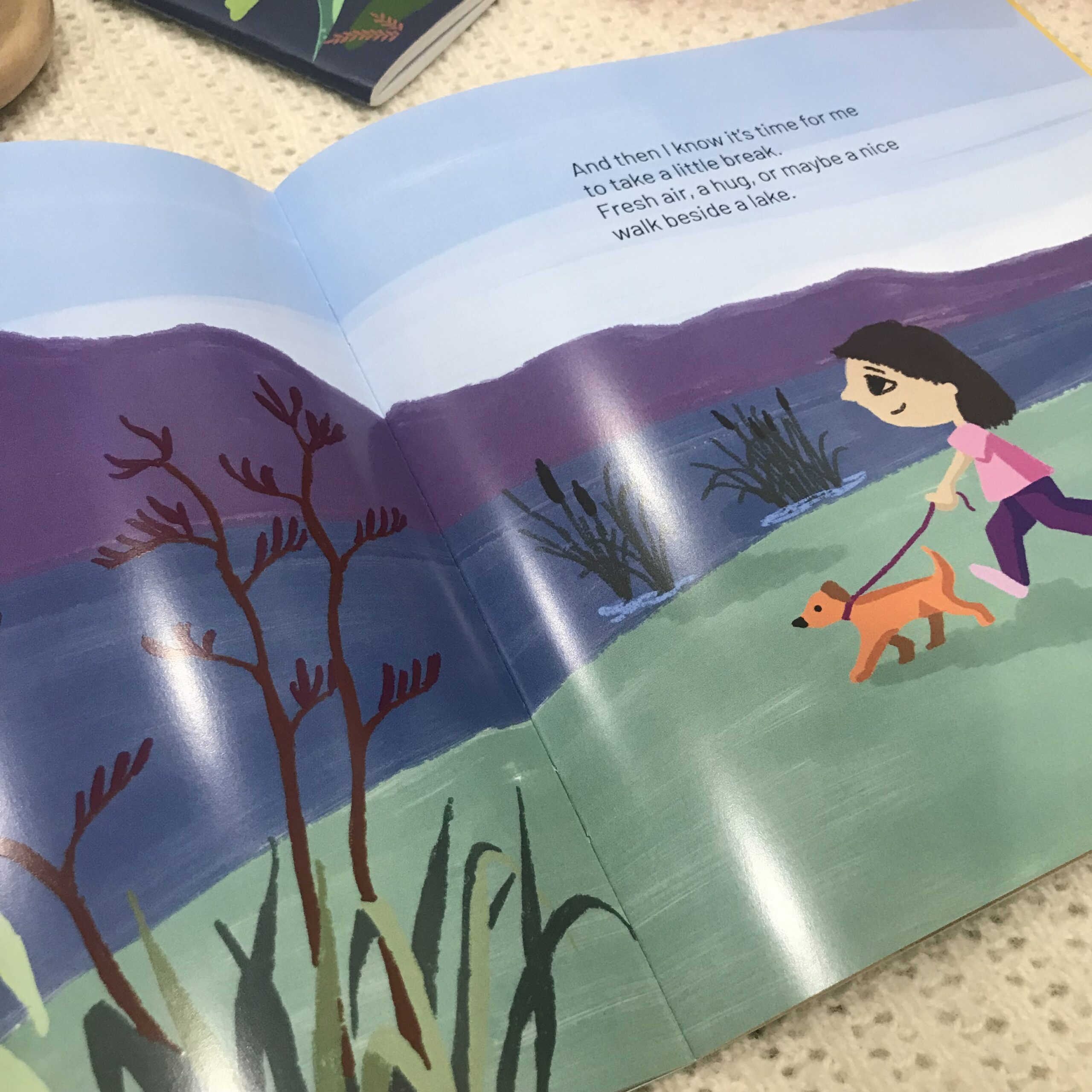 Picture of children's story book, There's a T-Rex in my Tummy" laid open with two pages showing. The pages show a small girl walking a dog beside a lake.